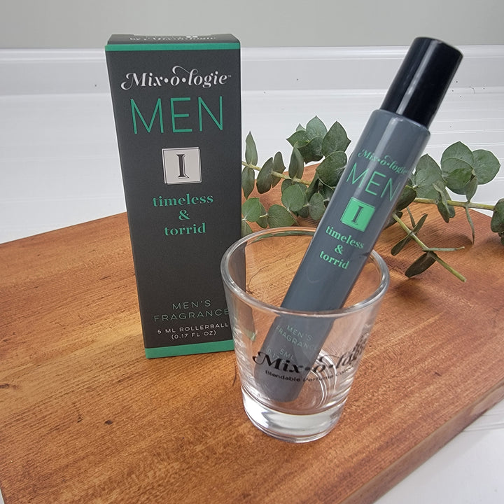 A Mixologie Men’s Fragrance rollerball rests in a branded glass beside its green packaging, with greenery in the background on a light surface.