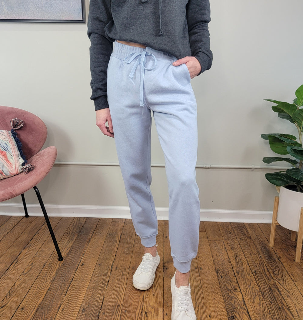 A person stands indoors wearing Rae Mode's Ember French Terry Sweatpants in light blue, paired with a dark gray cropped hoodie and white sneakers. One hand is in their pocket, surrounded by cozy decor like a pink chair, plant, and wood flooring.
