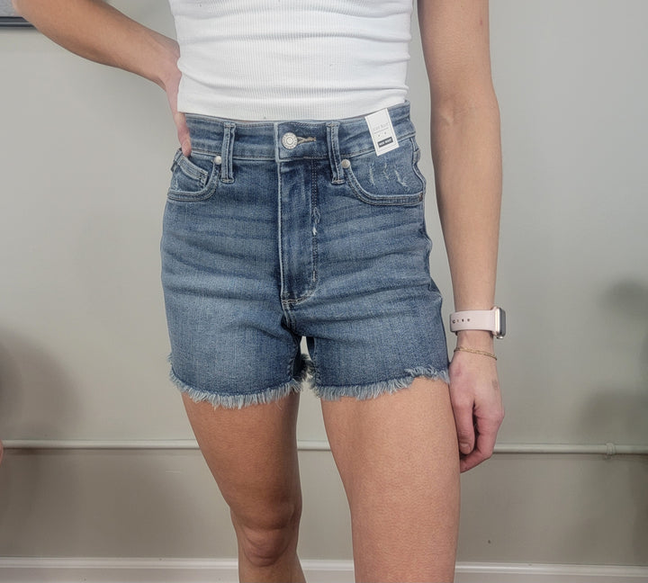 A person in a white ribbed tank top and Judy Blue Madeline Tummy Control Shorts stands against a neutral wall, left hand on their hip, wearing a smartwatch on their right wrist.