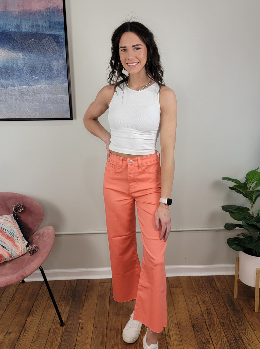 A smiling woman stands indoors wearing Zenana's Callie Colored Cropped Flares in coral and a white sleeveless top, beside a pink chair with a pillow, green plant, and framed art on a cream wall.