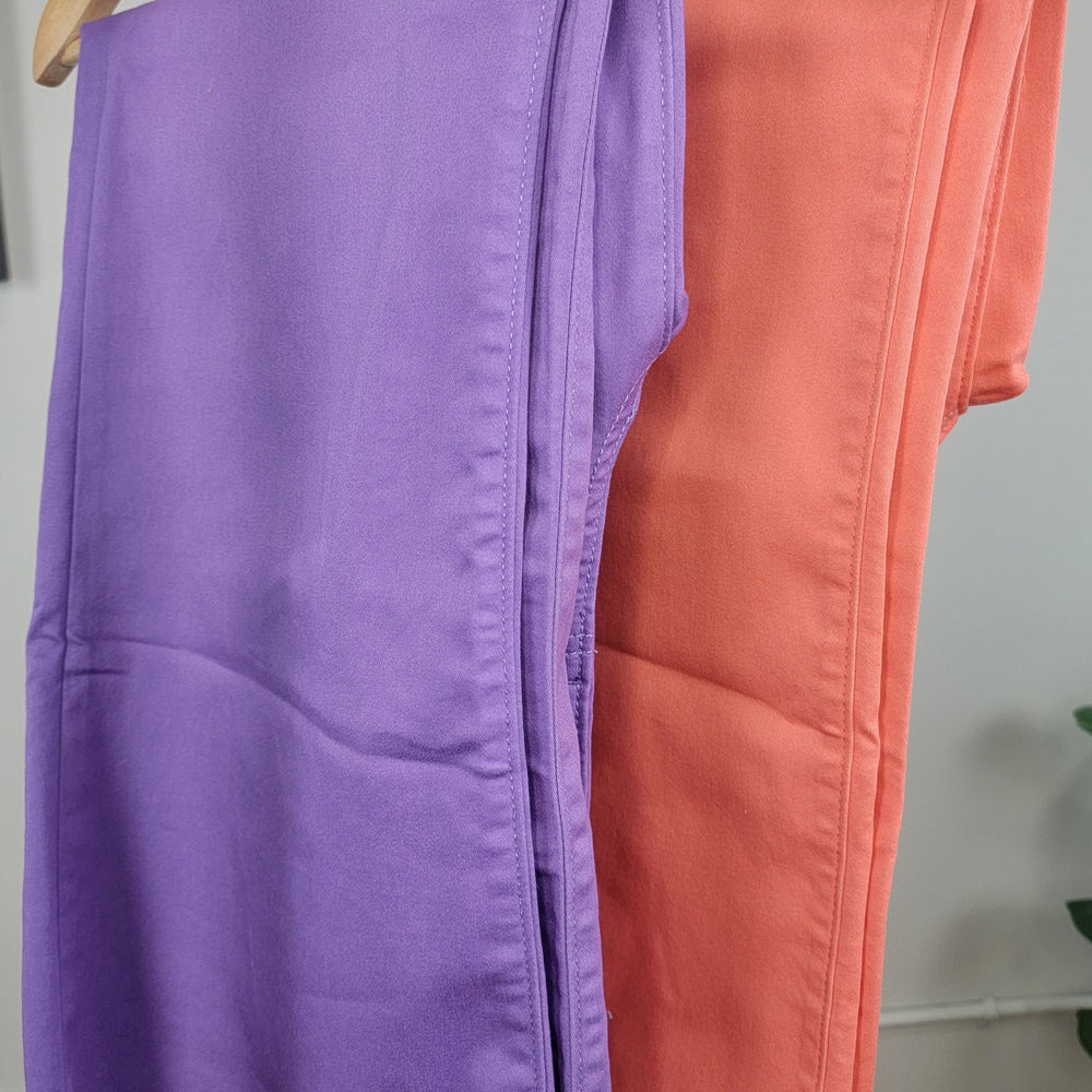 Two pairs of Zenana Callie Colored Cropped Flares hang side by side on hangers, one in bright purple and the other in orange, with a green plant partially visible in the background.