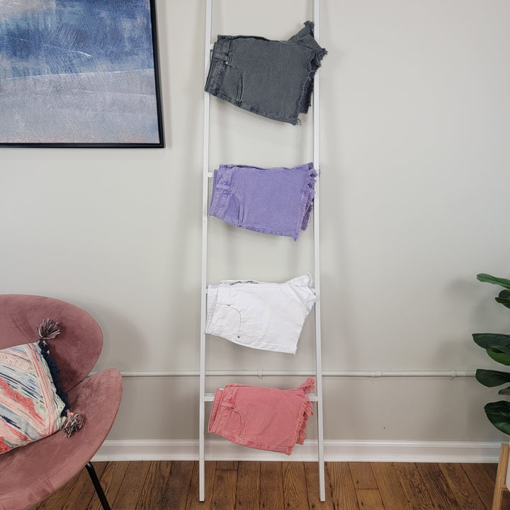 A white ladder leans against a wall, showcasing four folded pairs of Zenana Valeria Acid Wash Shorts in gray, purple, white, and pink. Nearby are a pink chair with a patterned pillow and a green plant on wooden flooring.