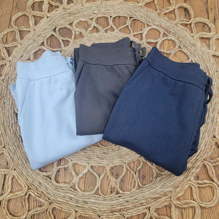 Three pairs of Rae Mode Ember French Terry Sweatpants in light blue, charcoal gray, and navy blue are folded side by side on a woven round mat on a wooden floor.