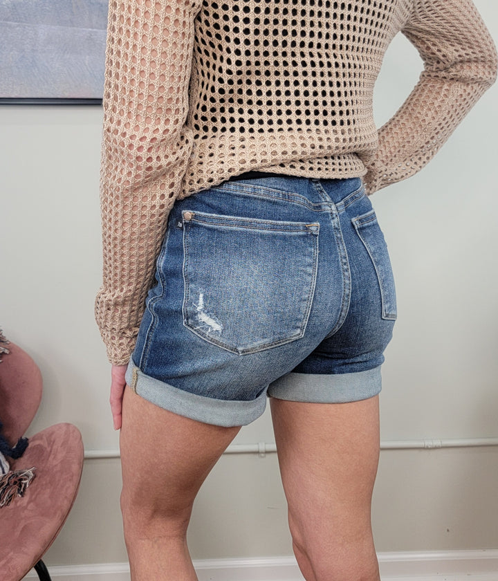A person wearing Siera Cuffed Shorts from Judy Blue and a beige, long-sleeve mesh-knit top stands facing away from the camera in a minimal, light-walled room.