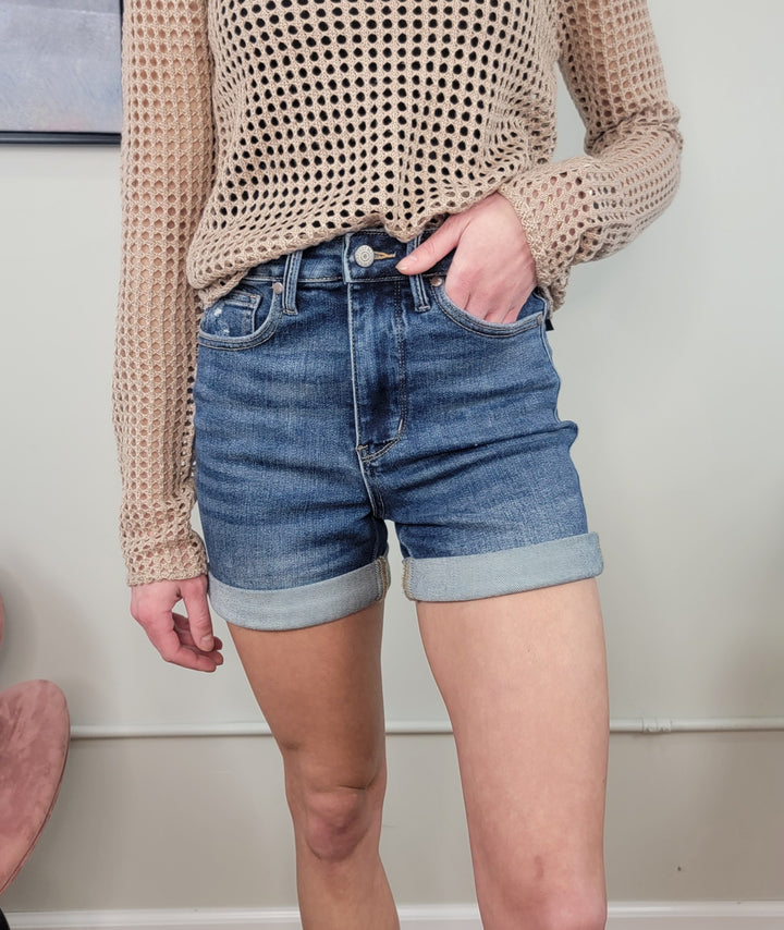 A person in a beige long-sleeve open-knit sweater and Judy Blue Siera Cuffed Shorts stands indoors with one hand in a pocket; their face is not shown.