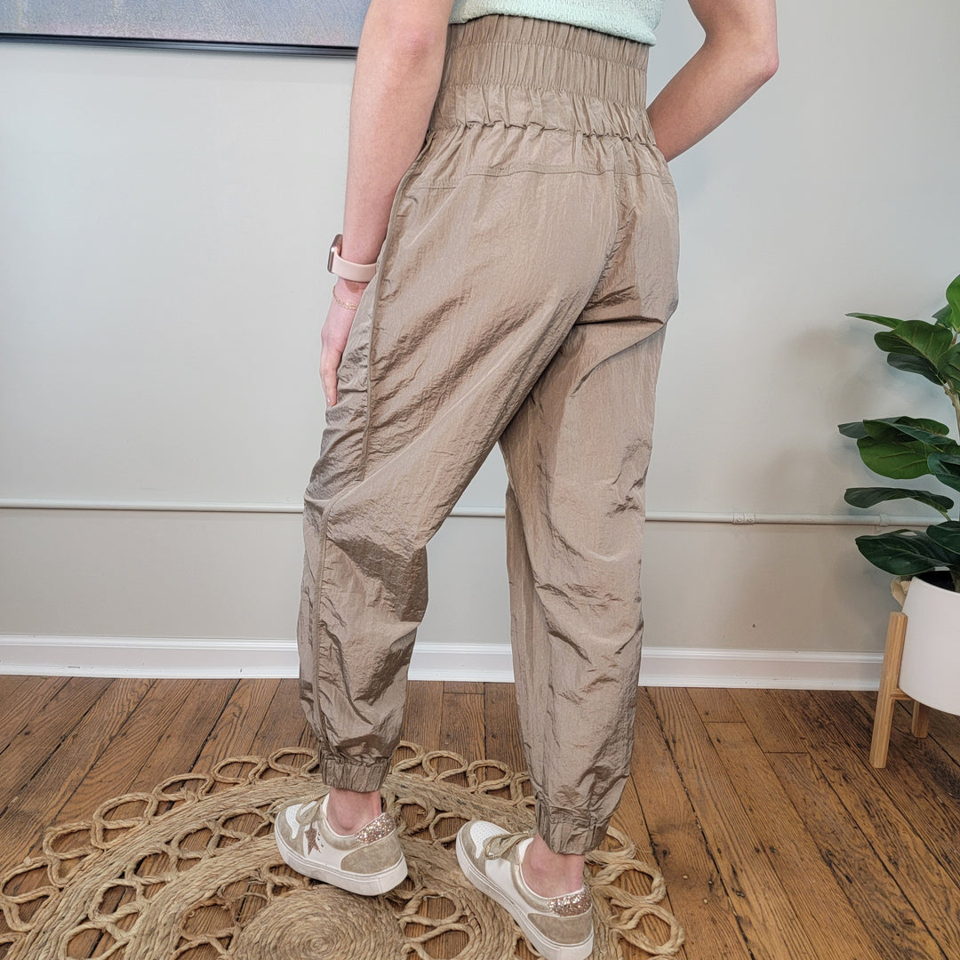 A person stands indoors on a circular woven rug, wearing GiGio Monroe Joggers with a smocked waistband and beige sneakers. Part of a green top is visible, with a plant and painting in the background.
