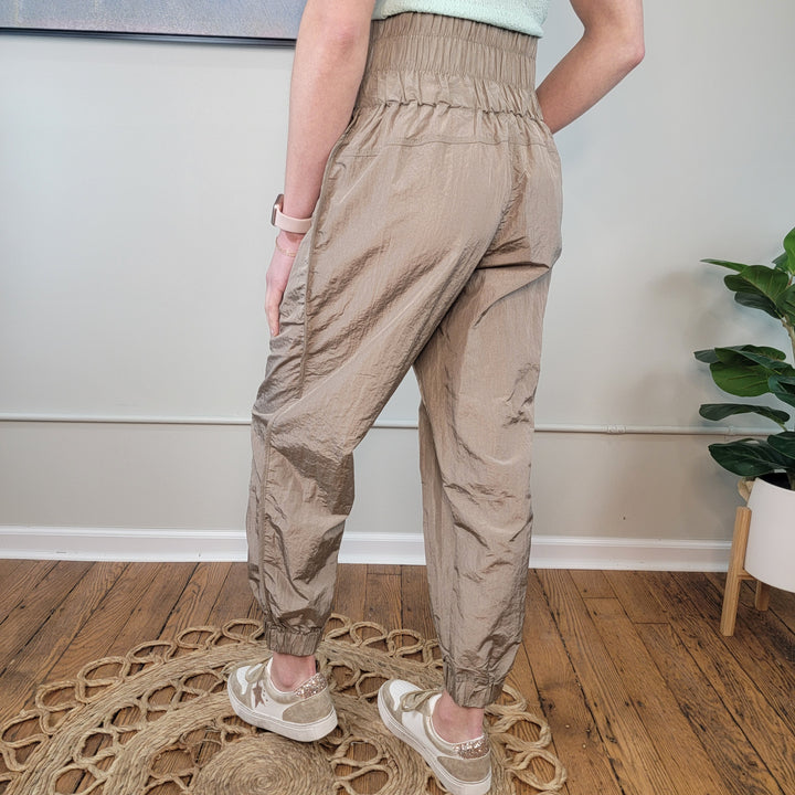 A person stands indoors on a circular woven rug, wearing GiGio Monroe Joggers with a smocked waistband and beige sneakers. Part of a green top is visible, with a plant and painting in the background.