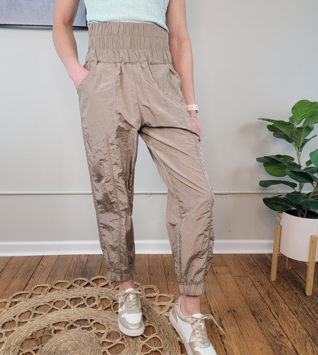 A person wearing GiGio Monroe Joggers in beige, a light green sleeveless top, and beige sneakers stands on a wooden floor by a round woven rug and a potted plant against a light-colored wall.