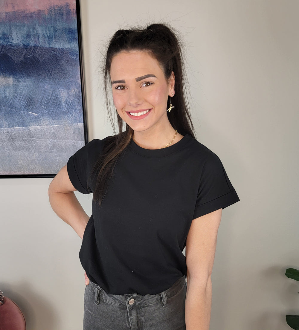 A woman with long dark hair styled half-up smiles indoors, wearing a Zenana Sibyl Cotton Tee in black and gray jeans, one hand on her hip. Behind her hangs a colorful abstract painting on the wall.