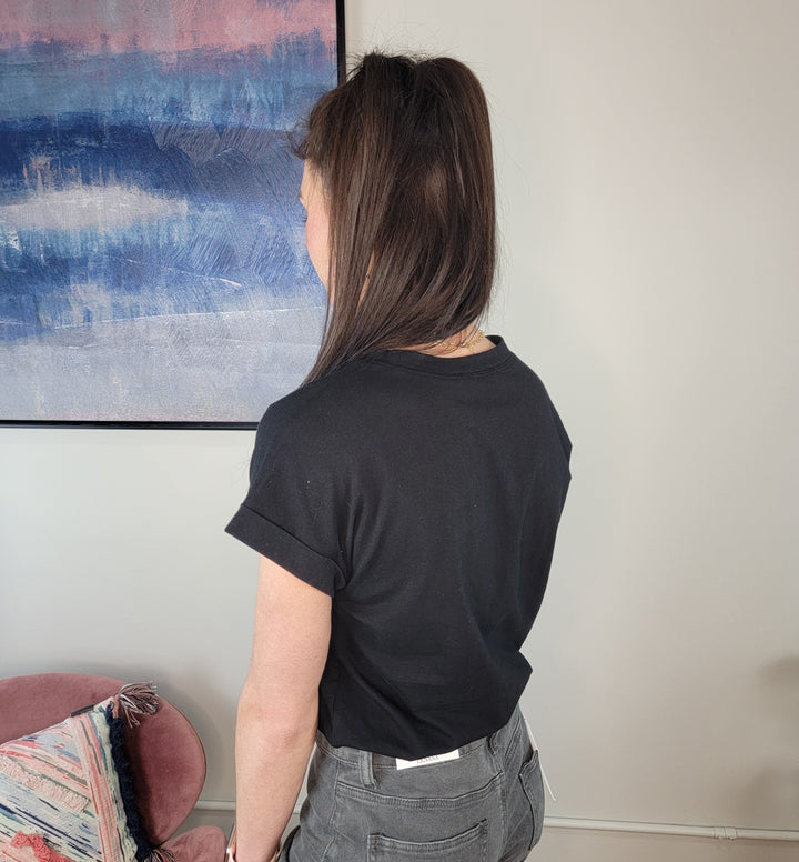 A person with straight brown hair wears a relaxed fit Sibyl Cotton Tee by Zenana and gray pants, standing with their back to the camera in a room with a pink chair and a blue and pink abstract painting on the wall.