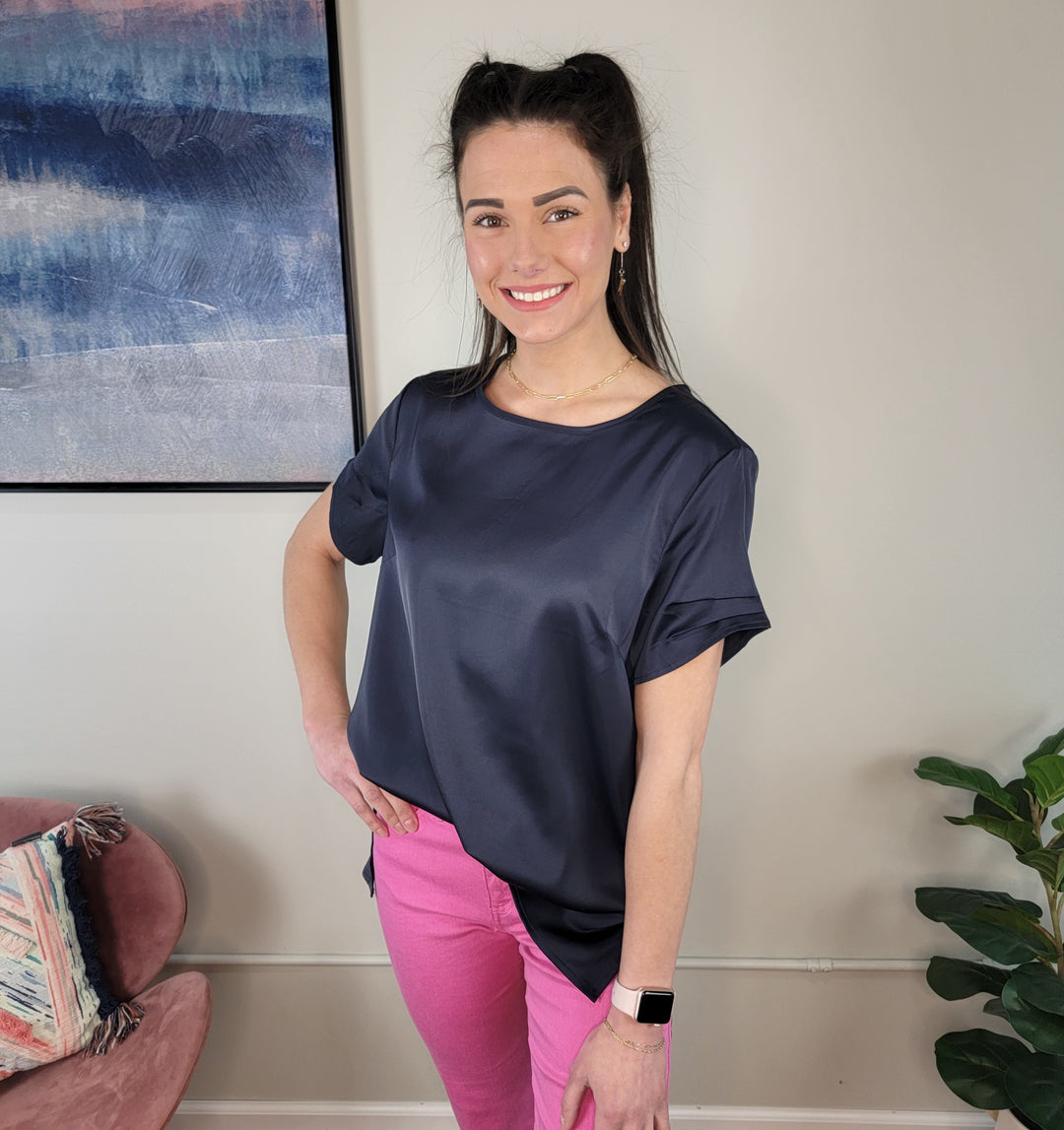 A woman with long dark hair in a half-up hairstyle smiles at the camera, wearing the GiGio Greta Top in navy, paired with bright pink pants and a pink smartwatch. She stands indoors near a plant and abstract wall art.