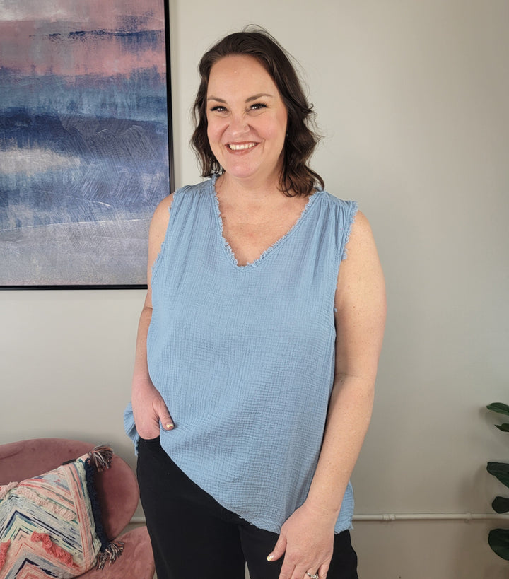 A woman with shoulder-length brown hair wears the Umgee Jennifer Sleeveless Top in light blue and black pants, smiling at the camera indoors beside a framed abstract painting and a pink chair with a patterned cushion.
