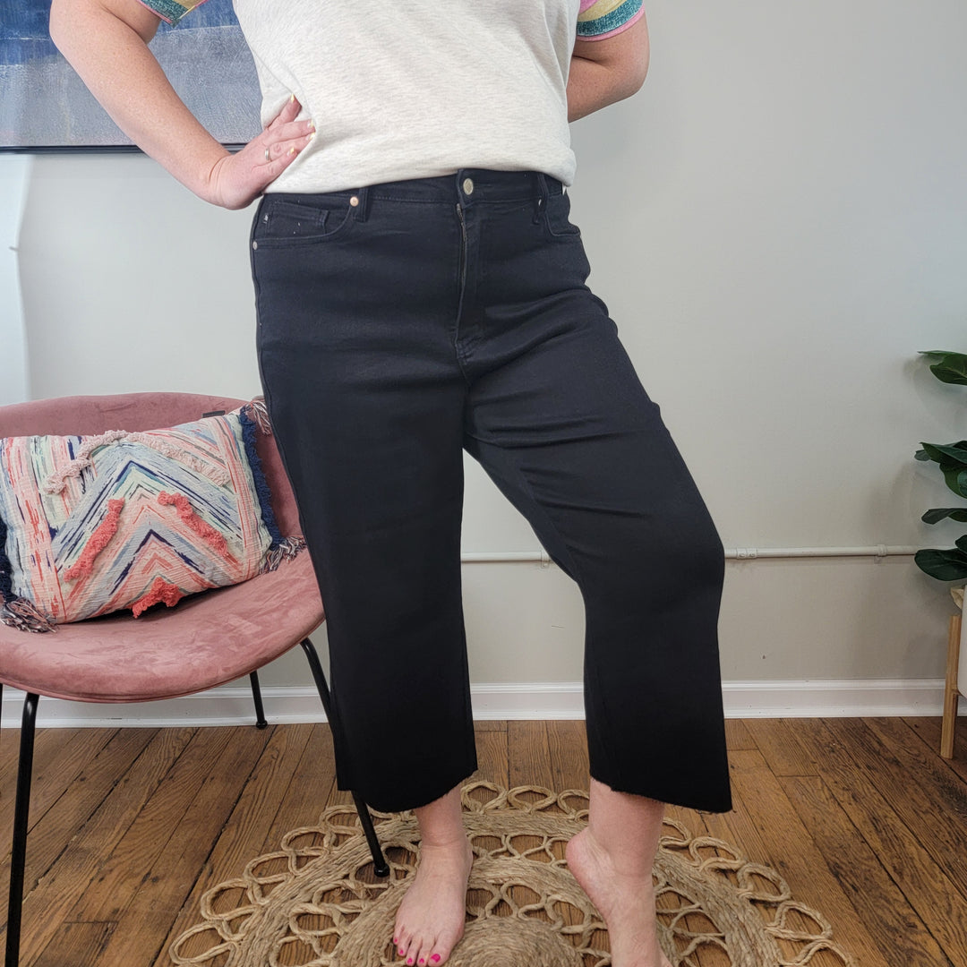 A person wearing Judy Blue's Rylee Tummy Control Cropped Wide Legs stands barefoot on a wooden floor beside a pink chair with a colorful pillow and a small green plant.