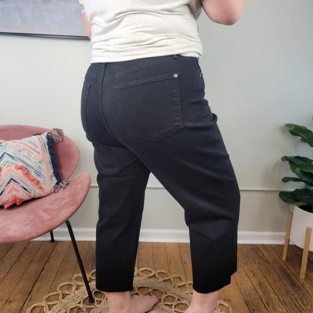 A person wearing the Rylee Tummy Control Cropped Wide Legs jeans from Judy Blue stands barefoot on a woven rug in a room with wooden floors, a pink chair, a colorful pillow, and a green plant in the background.
