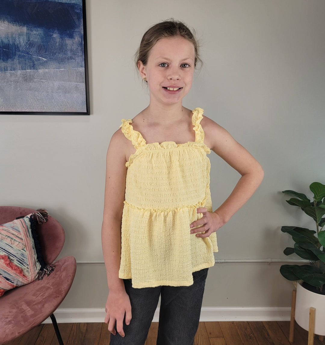 A young girl with light brown hair stands indoors, wearing the Hayden Talulla Tank Top in yellow and dark pants. Behind her are a pink chair with a colorful pillow, a green plant, and framed wall art.