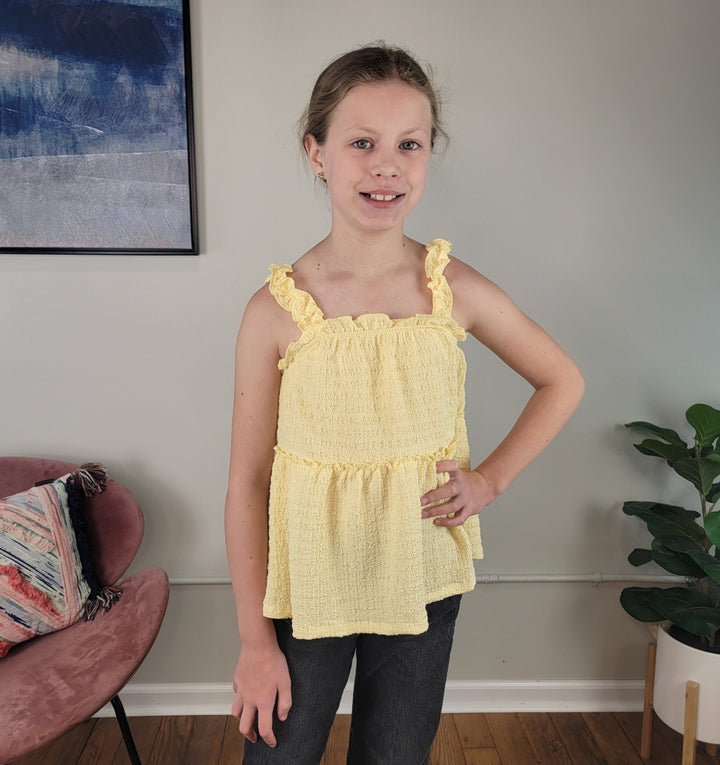 A young girl with light brown hair stands indoors, wearing the Hayden Talulla Tank Top in yellow and dark pants. Behind her are a pink chair with a colorful pillow, a green plant, and framed wall art.