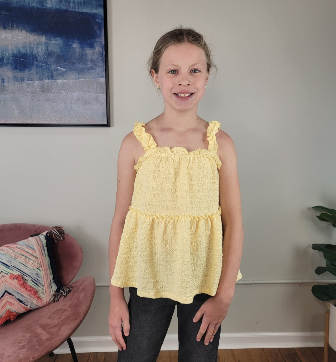 A smiling girl with light brown hair in a ponytail wears the Hayden Girls: Talulla Tank Top, standing indoors near a pink chair, a plant, and a blue abstract painting on a beige wall.
