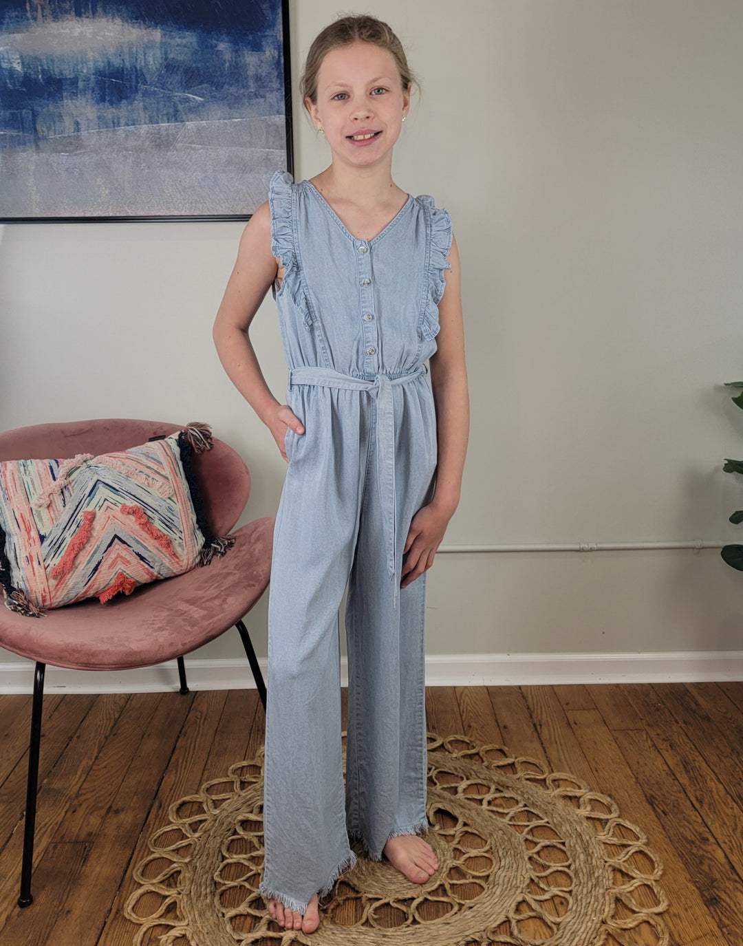 A girl wears the Hayden Girls: Rylan Jumper, a sleeveless 100% Tencel chambray piece. She stands barefoot on a woven rug, smiling at the camera with one hand in her pocket. A pink chair with a colorful pillow and a plant are behind her.