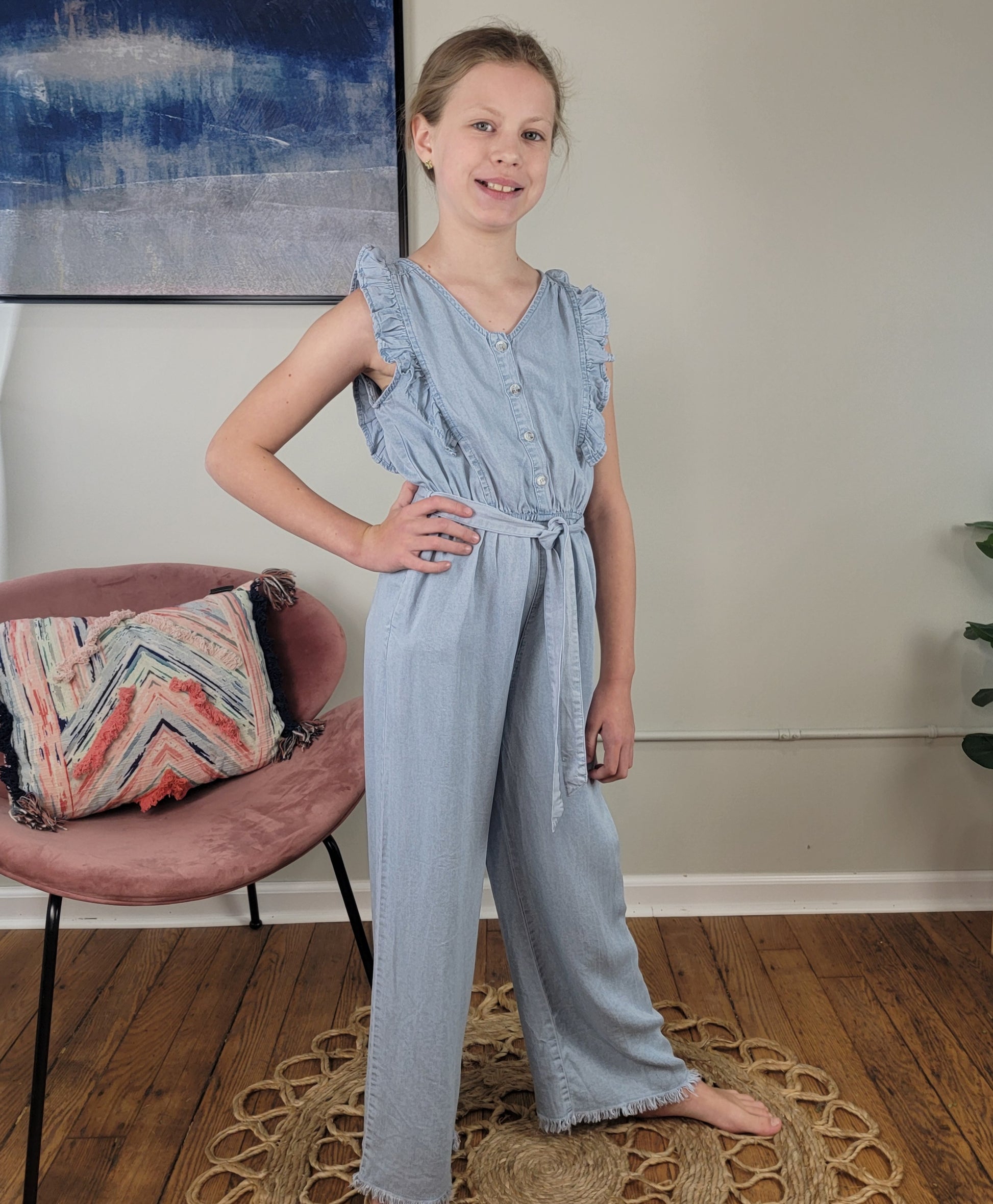 A young girl poses confidently indoors on a round rug, smiling in the Hayden Girls: Rylan Jumper—a light blue Tencel sleeveless romper with ruffle details and a tie belt. A pink chair and abstract painting are in the background.
