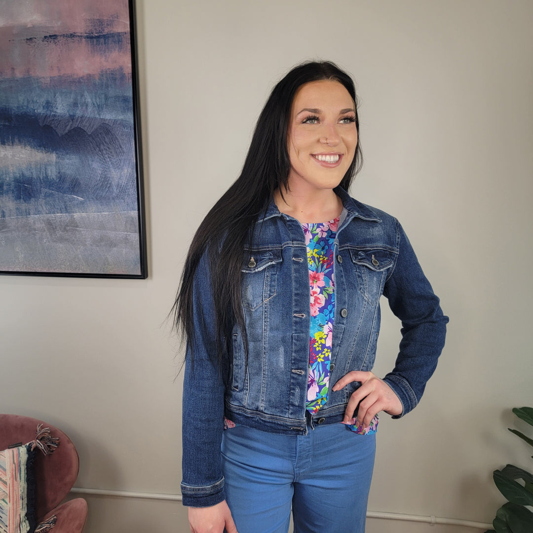 A woman with long dark hair smiles indoors, wearing the Hollis Denim Jacket by Risen over a colorful floral top and blue pants. A modern abstract painting hangs on the wall behind her.