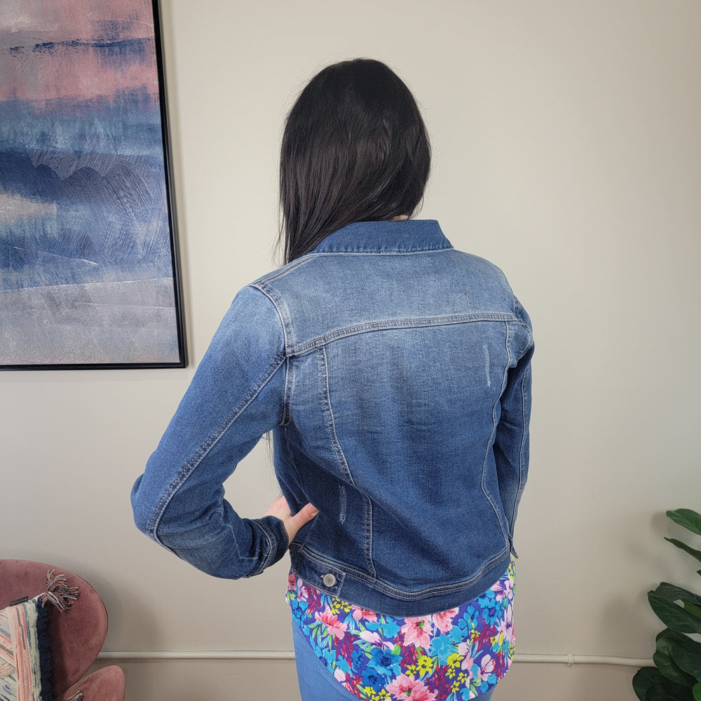 A woman with long dark hair stands indoors facing away, wearing the Risen Hollis Denim Jacket over a colorful floral top. The room has light walls, a framed abstract painting, a pink chair, and a green plant.
