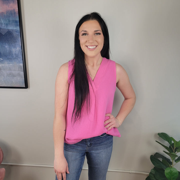 A woman with long dark hair wears the Staccato Kendra Blouse, a sleeveless v-neck top, paired with blue jeans as she stands and smiles in a room featuring neutral walls, a framed picture, and a green plant in the corner.
