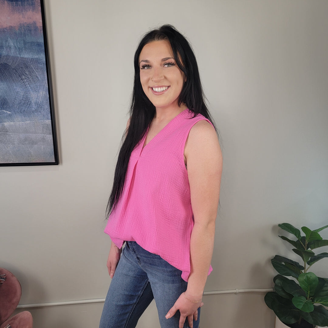 A woman with long dark hair smiles indoors, wearing the Staccato Kendra Blouse—a sleeveless v-neck top in pink—paired with blue jeans. A framed picture hangs on the wall and a green potted plant sits nearby.
