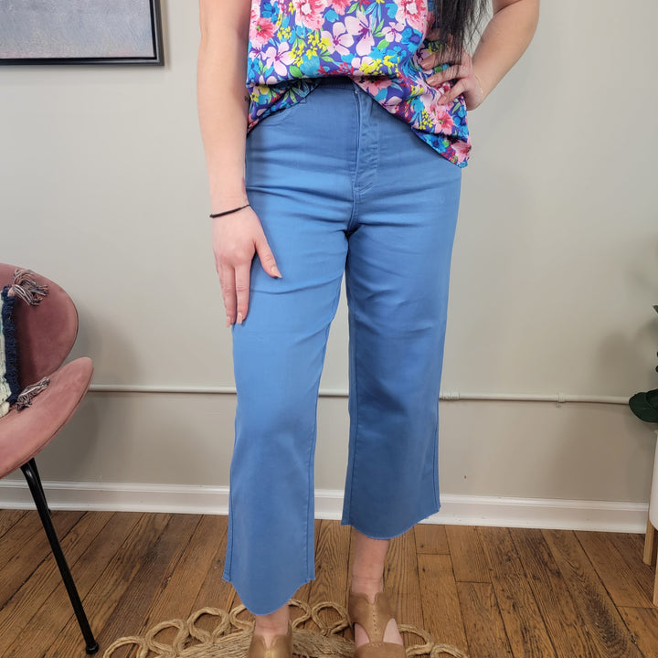 A person stands indoors on a wooden floor, wearing Judy Blue’s Elexis Tummy Control Wide Leg Crops in light blue, paired with a colorful floral top and tan sandals. One hand rests on their thigh and the other is on their hip.