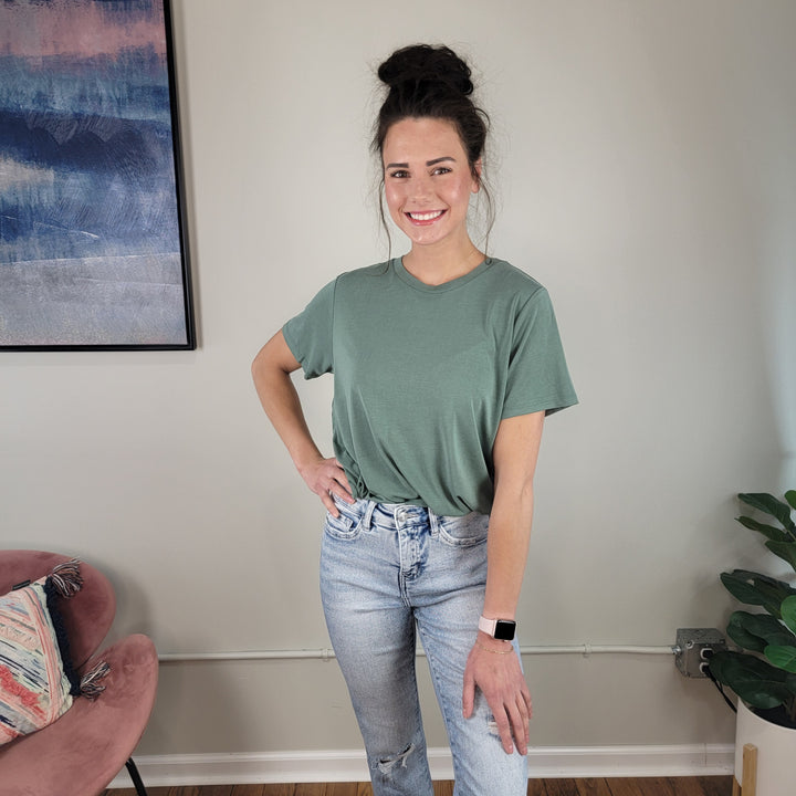 A woman with dark hair in a bun smiles with one hand on her hip, wearing the Keely Tee by HYFVE, Inc and light blue jeans, standing in a modern room with a plant, pink chair, and abstract wall art.