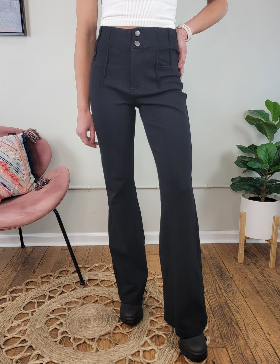 A person stands indoors on a wooden floor next to a pink chair and green potted plant, wearing Rae Mode's Landry Stretch Dress Pants—high-waisted, black, with a flare leg—paired with a white sleeveless top.