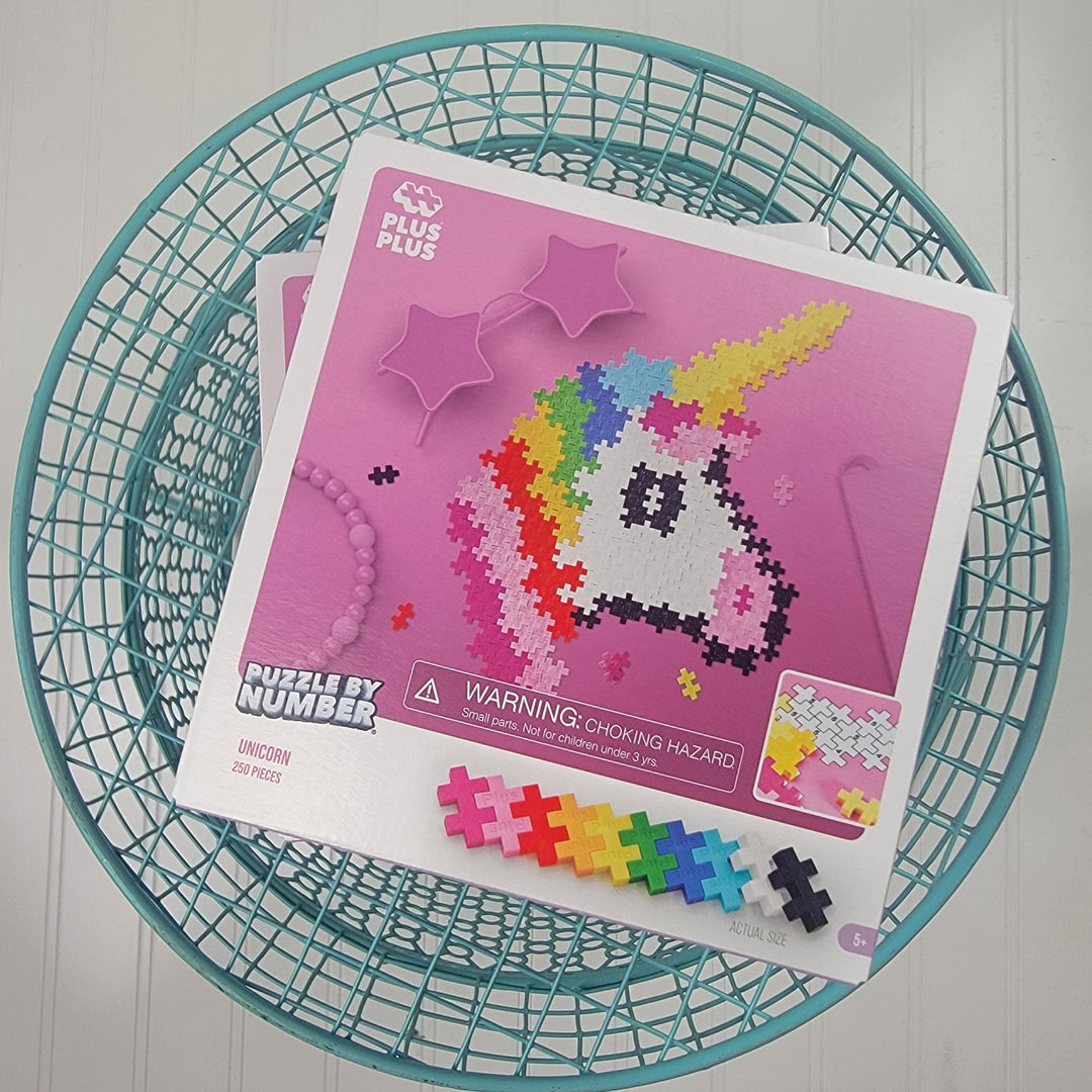 The Plus Plus Puzzle By Number Unicorn kit shows a pixelated unicorn design on the box, colorful pieces below, and is placed in a turquoise wire basket on a white surface.