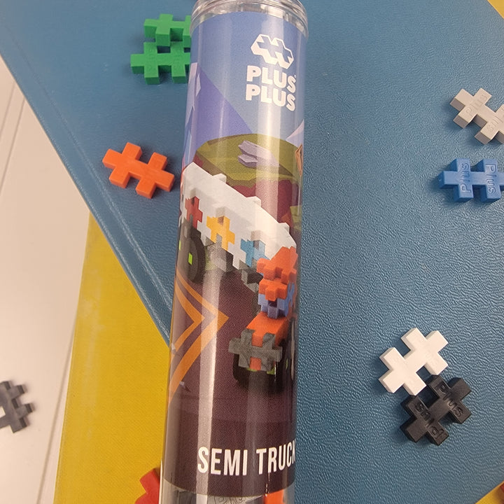 A Plus Plus Puzzle Tubes toy from Plus Plus rests on a blue surface, surrounded by colorful building pieces. The tube displays an image of a semi truck built with these unique construction toys.
