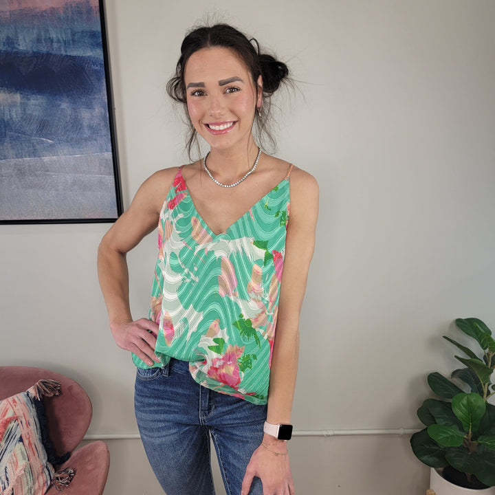 A woman with dark hair in a loose updo smiles at the camera, wearing the Staccato Tyra Floral Tank with adjustable straps, blue jeans, a pearl necklace, and a smartwatch. She stands indoors near a plant and abstract wall art.