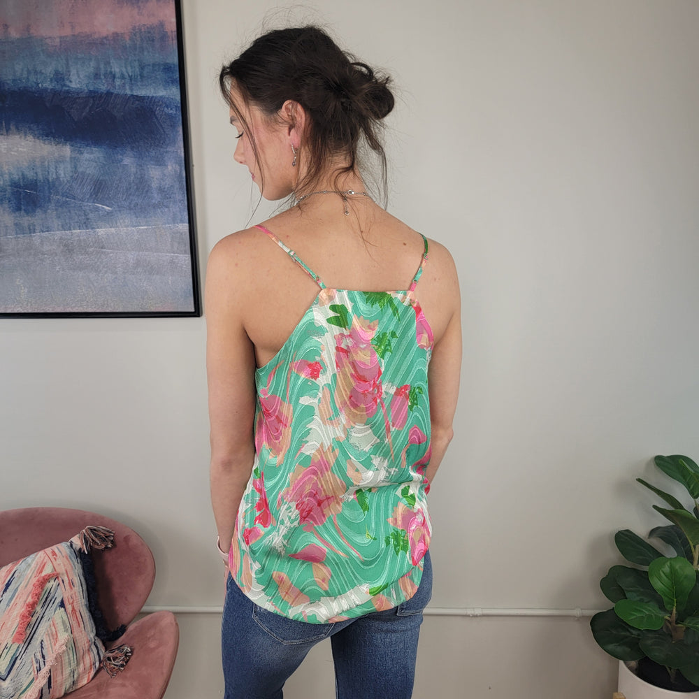 A woman stands with her back to the camera, wearing the Staccato Tyra Floral Tank with adjustable straps and blue jeans in a room featuring a pink chair, a plant, and abstract wall art.