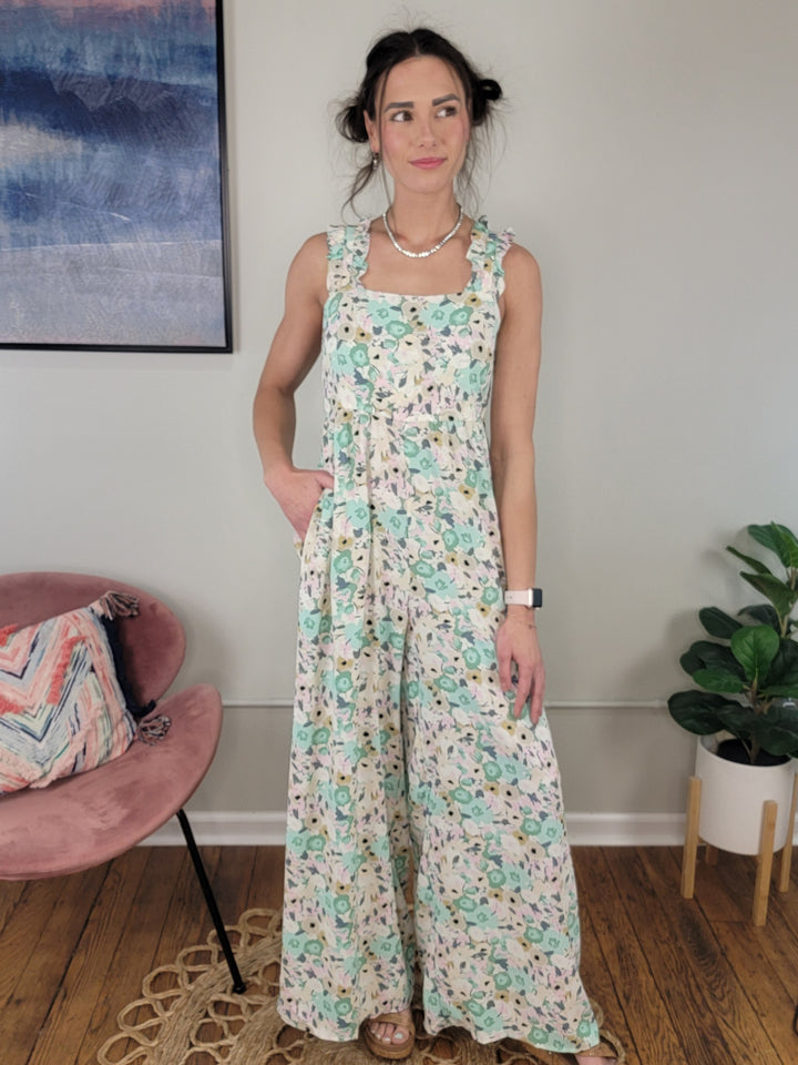 A woman indoors wears the Kori America Milani Floral Jumpsuit, a sleeveless sage piece with ruffle straps and a back tie. She styles her hair in two buns with accessories. The room includes a pink chair, plant, and wall art.