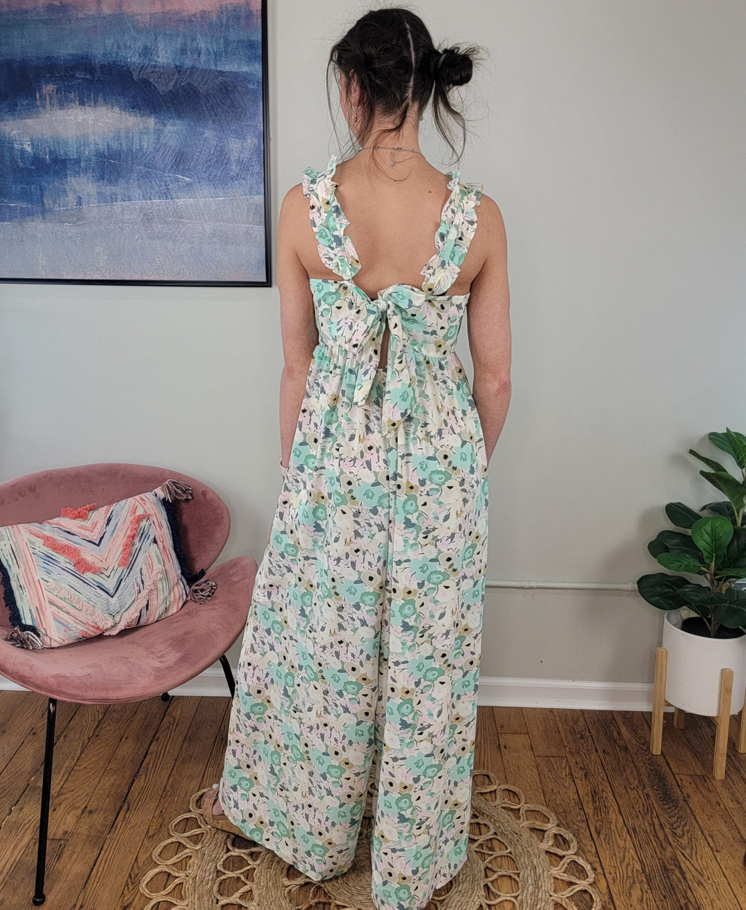 A woman with dark hair in a messy bun wears the Milani Floral Jumpsuit by Kori America, featuring ruffle straps and a back tie. She stands on a rug, facing away, in a room with a pink chair, plant, and framed painting.
