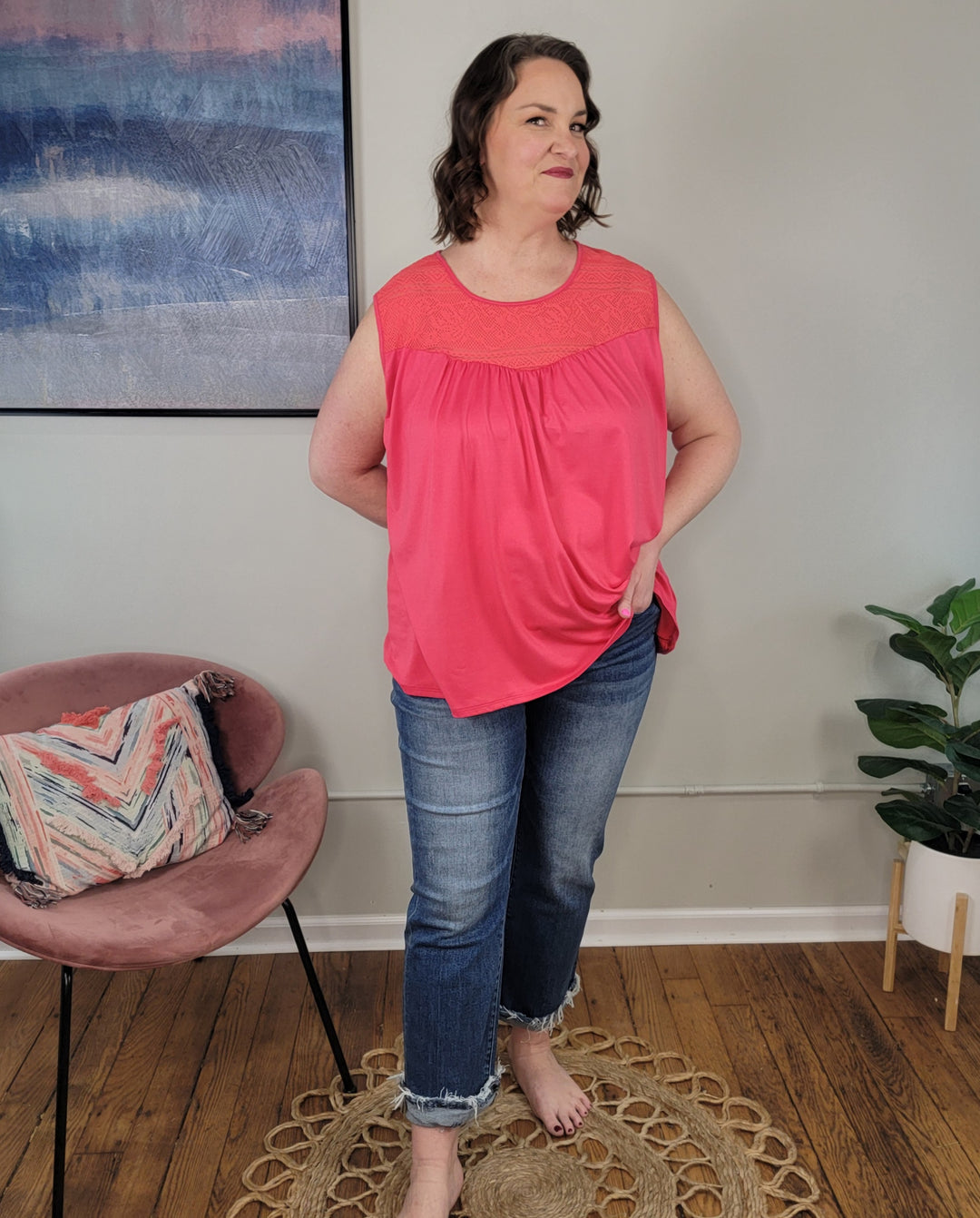 A woman stands indoors on a circular rug, wearing the Haptics Adira Sleeveless Top in coral and cuffed blue jeans. She smiles with one hand in her pocket; behind her are a pink chair with cushion, a plant, and framed artwork on the wall.