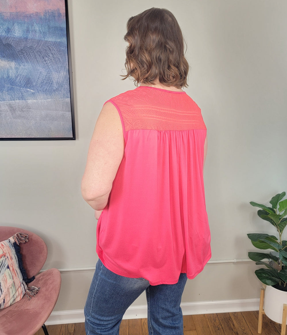 A person with wavy brown hair faces away indoors, wearing the Haptics Adira Sleeveless Top in coral and blue jeans. The room features a plant, pink chair, and framed artwork on the wall.