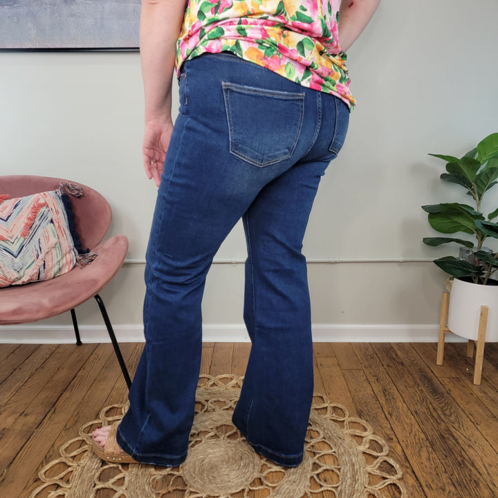 A person in Risen's Pennie Bootcut Jeans faces away from the camera, standing on a circular woven rug in a room with wooden floors, a pink chair, and a potted plant.