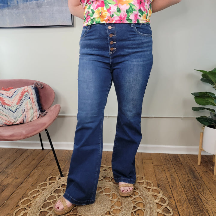 A person stands indoors on a wooden floor near a chair with a patterned pillow and potted plant, wearing Pennie Bootcut Jeans from Risen paired with a colorful floral top featuring large buttons.