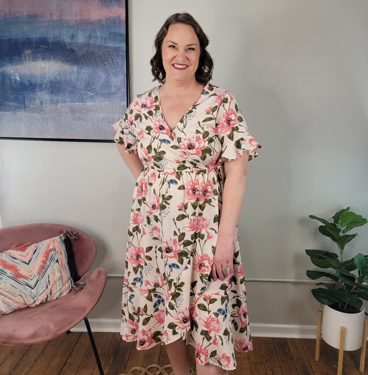 A woman wearing the Kori America Ilaria Floral Midi Dress with ruffle detail stands indoors by a pink chair, a colorful pillow, and a potted plant. Behind her hangs a blue and pink abstract painting.