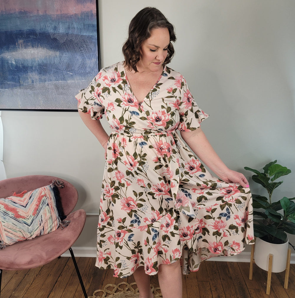 A woman wears the Kori America Ilaria Floral Midi Dress with ruffle detail, standing indoors as she holds her skirt. Nearby are a pink chair with a pillow, a green potted plant, and a blue and pink abstract painting.