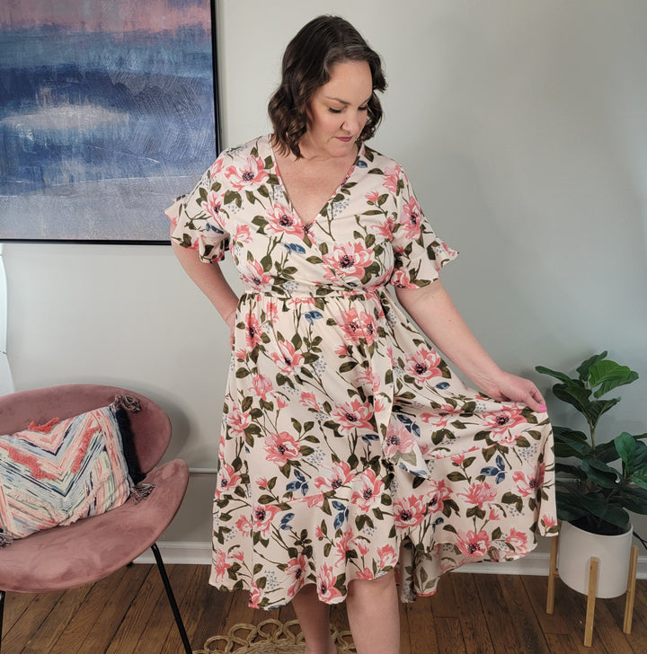 A woman wears the Kori America Ilaria Floral Midi Dress with ruffle detail, standing indoors as she holds her skirt. Nearby are a pink chair with a pillow, a green potted plant, and a blue and pink abstract painting.