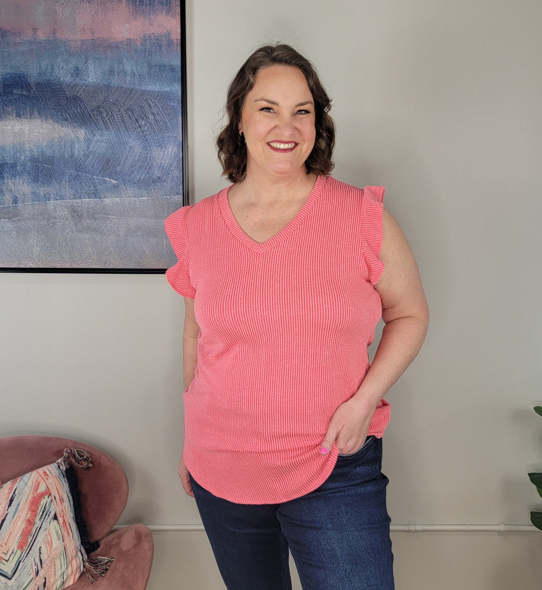 A woman with short brown hair smiles indoors, wearing the Heimish Trixie Textured Top in coral and dark jeans. She stands with one hand in her pocket near a pink chair, a patterned pillow, and a large abstract painting on the wall.