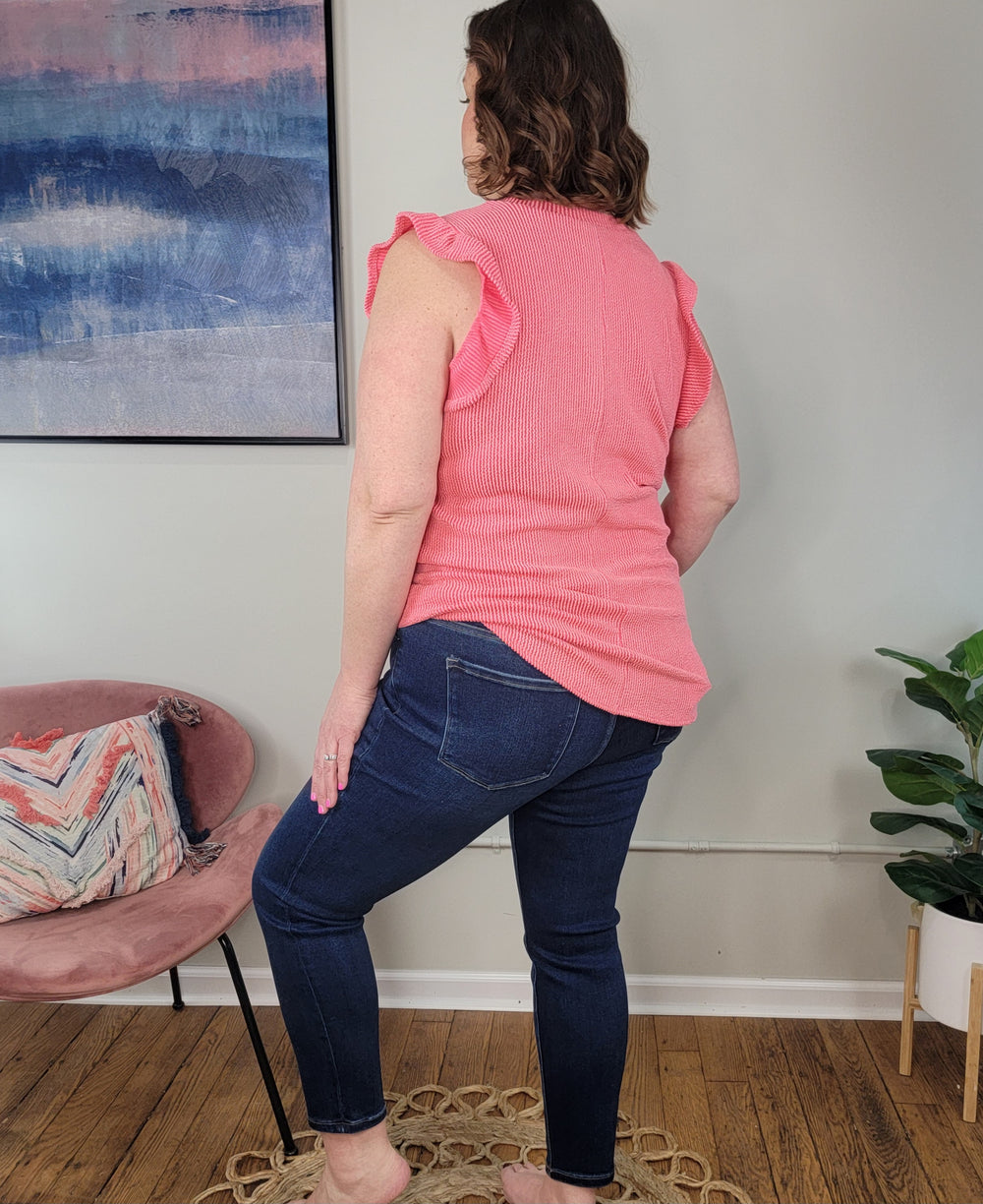 A woman stands indoors with her back to the camera, wearing the Heimish Trixie Textured Top in coral and dark blue jeans. She is beside a pink chair with a decorative pillow, a plant, and a painting on the wall.