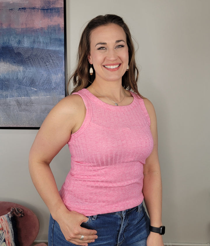 A woman with long brown hair smiles indoors, wearing the Zenana Bridget Tank Top in pink and denim shorts—ideal for summer layering. An abstract painting decorates the wall, and a chair draped with a blanket sits in the background.