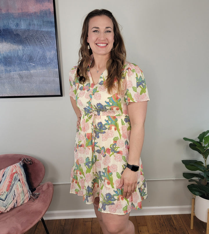 A woman in the Doe & Rae Lark Dress stands indoors, smiling at the camera. She wears a black smartwatch; behind her are a pink chair, a plant, and abstract wall art.