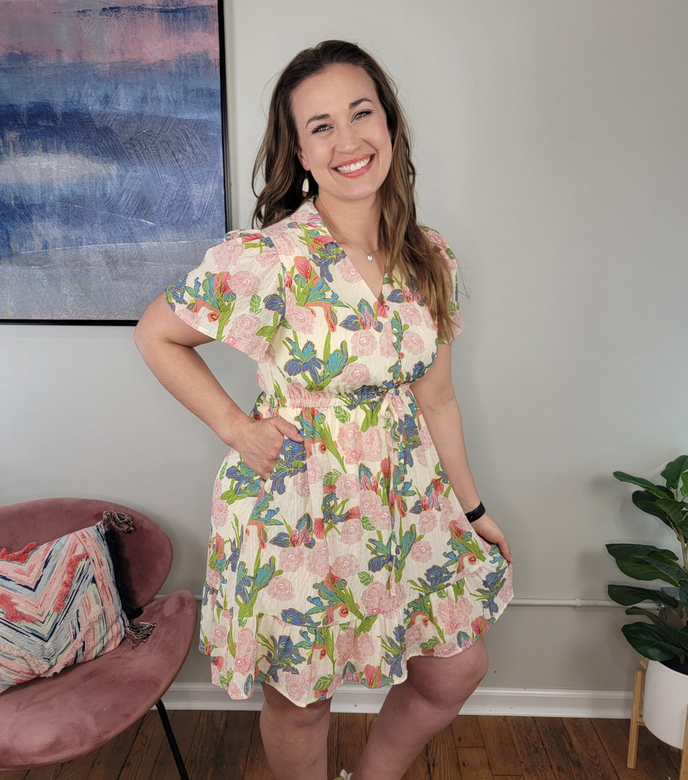 A woman stands gracefully in the Lark Dress by Doe & Rae, its floral print and ruffle hem adding a touch of elegance to her look.