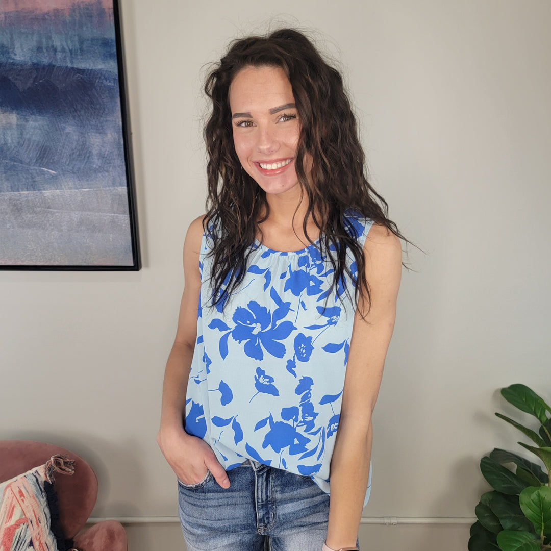 A smiling woman with wavy brown hair stands indoors near a green plant, wearing jeans and the Staccato Andaz Floral Tank, a lightweight two-tone blue floral top with pleating details. A framed picture hangs on the wall behind her.