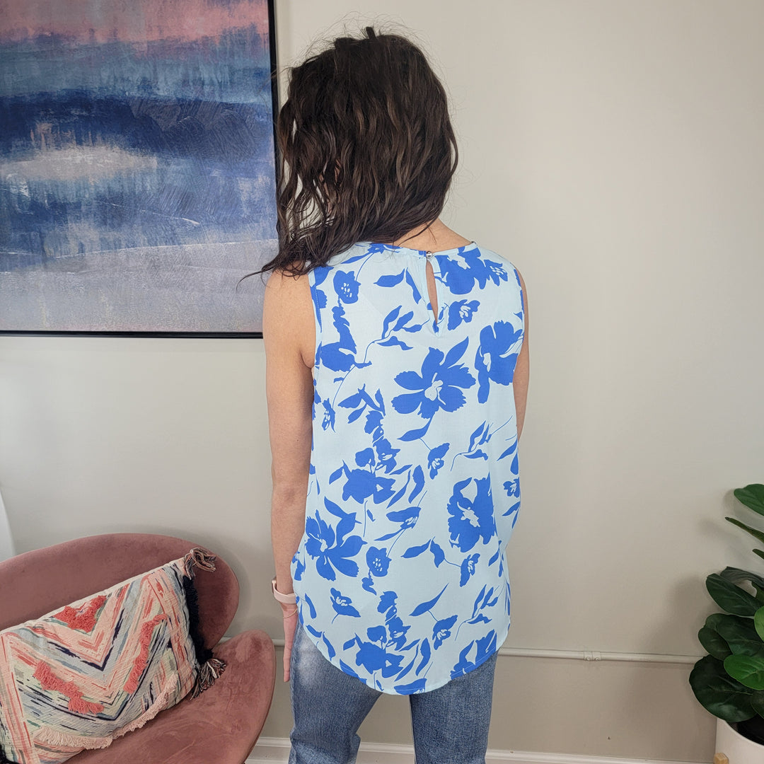 A person with wavy brown hair wears the Staccato Andaz Floral Tank and blue jeans, standing with their back to the camera in a room featuring a pink chair, colorful pillow, and large abstract wall art.